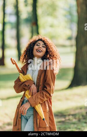 Donna entusiasta in impermeabile guardando la macchina fotografica mentre tiene l'ombrello nel parco Foto Stock