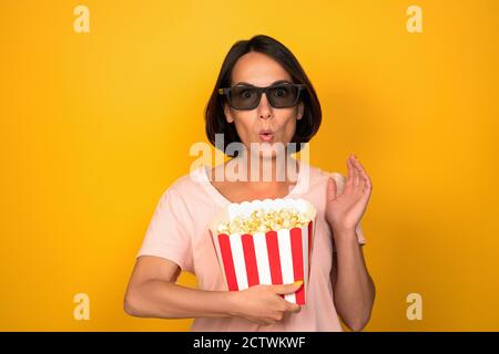 Donna in bicchieri scuri contiene scatola di popcorn. Giovane brunetta con uno sguardo sorpreso alzò la mano. Ritagliato su sfondo giallo in studio. Cinema Foto Stock