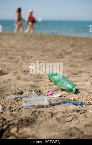 Verde, bottiglia di plastica, bastoncini di cotone, tappo, scatola e sigarette lasciati su una spiaggia sabbiosa con persone che camminano sullo sfondo. Foto Stock