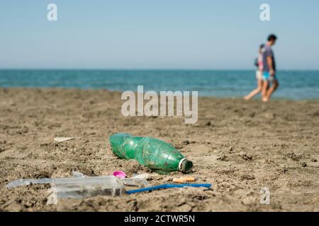 Verde, bottiglia di plastica, bastoncini di cotone, tappo, scatola e sigarette lasciati su una spiaggia sabbiosa con persone che camminano sullo sfondo. Foto Stock