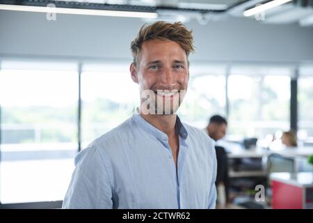 Ritratto di sicuro caucasico maschile business creativo con capelli corti e barba sorridente, con colleghi che lavorano in background. Business creativo Foto Stock