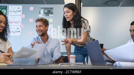 Donna d'affari asiatica in piedi per affrontare sorridenti colleghi di sesso maschile e femminile in possesso di documenti durante una riunione di squadra. I professionisti creativi di affari lo faranno Foto Stock