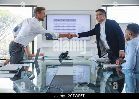 Uomo d'affari asiatico e caucasico in piedi scuotendo le mani sul tavolo in una riunione di affari, con i colleghi seduti accanto a loro. Business creativo Foto Stock