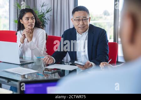 Uomo d'affari asiatico anziano che tiene il tablet e parla durante una riunione di affari, una collega asiatica seduta accanto a lui che ascolta. Busine creativo Foto Stock