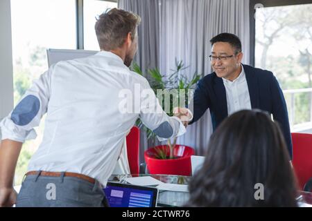Uomo d'affari asiatico e caucasico in piedi scuotendo le mani sul tavolo in una riunione di affari, con i colleghi seduti accanto a loro. Business creativo Foto Stock