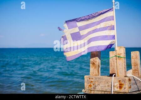 Un'antica bandiera greca strappata svetta nel vento, posta su una piccola sedia di legno, mare aperto in lontananza. Foto Stock