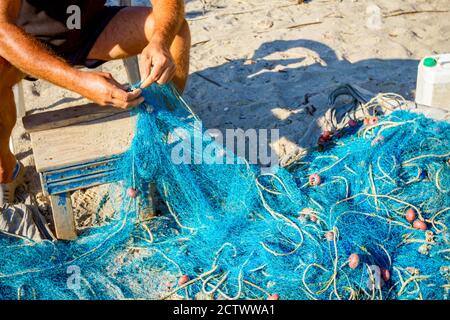 Un pescatore si siede su una sedia alla spiaggia, accumola la rete di pesca e la pulisce dalle conchiglie. Foto Stock