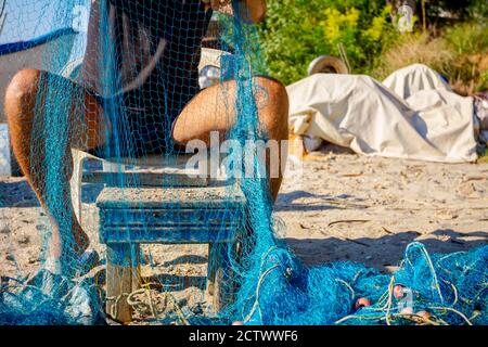 Un pescatore si siede su una sedia alla spiaggia, accumola la rete di pesca e la pulisce dalle conchiglie. Foto Stock