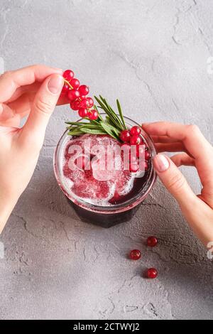 Le mani della donna contengono bacche di ribes per la preparazione di cocktail di frutta fresca ghiacciata in vetro con foglia di rosmarino, fondo di pietra in cemento Foto Stock