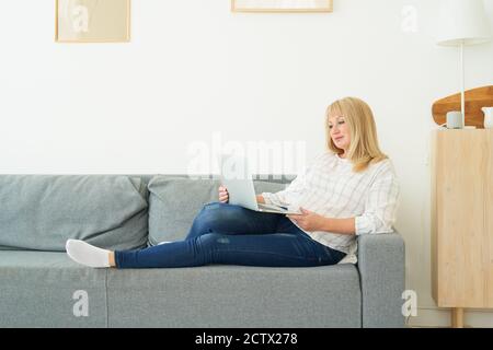 Donna matura a lunghezza intera che usa un computer portatile che si stesa su un comodo divano, interno di casa Foto Stock