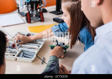 Fuoco selettivo dell'insegnante seduto vicino ai bambini che modellano il robot dentro in aula Foto Stock