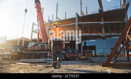 Back Shot del lavoratore appaltatore che indossa casco rigido e giubbotti di sicurezza cammina sul sito di costruzione di edifici industriali. In background Crane, Skyscraper Foto Stock