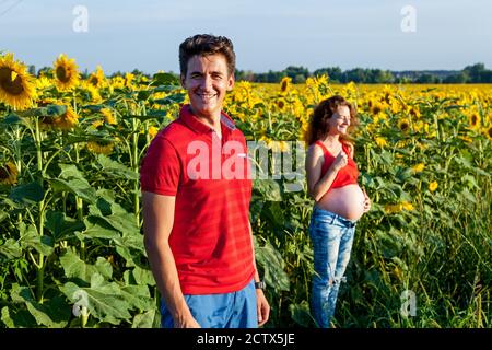 Un marito in piedi vicino alla sua moglie incinta nel campo con un sacco di girasoli in una giornata di sole. Foto Stock