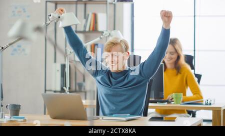Bell'esperto IT caucasico seduto alla sua scrivania lavora su un laptop, alza le braccia in Celebration dopo che ha finito con successo il progetto. Diversi Foto Stock