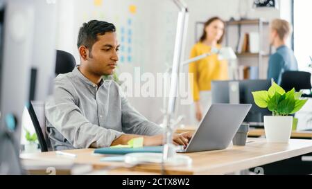 Un lavoratore indiano intelligente e bello seduto alla sua scrivania lavora su un laptop. In background Ufficio moderno con diversi team di giovani Foto Stock