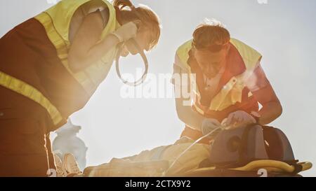 Sull'automobile Crash incidente di traffico scena: Paramedics che salva la vita di una vittima di incidente di traffico che sta mentendo su Stretchers. Ascoltano un Heartbeat Foto Stock