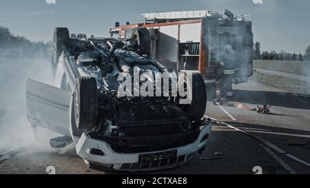 Incidente stradale ribaltamento veicolo e bruciare sdraiato sul suo tetto nel mezzo della strada dopo la collisione. Nel team di background di Foto Stock