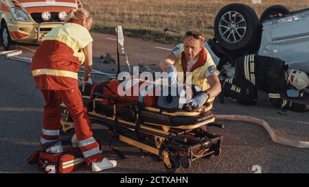 Sull'automobile Crash scena di incidente di traffico: Paramedics che salva la vita di una vittima femminile che sta mentendo su Stretchers. Applicano la maschera di ossigeno, fare Foto Stock