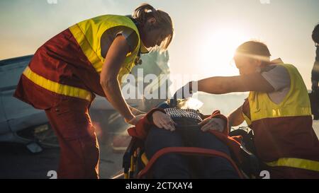 Sull'automobile Crash scena di incidente di traffico: Paramedics che salva la vita di una vittima femminile che sta mentendo su Stretchers. Ascoltano un Heartbeat, si applicano Foto Stock