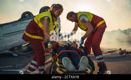 Sull'automobile Crash scena di incidente di traffico: Paramedics che salva la vita di una vittima femminile che sta mentendo su Stretchers. Ascoltano un Heartbeat, si applicano Foto Stock