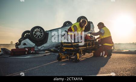 Sull'automobile Crash scena di incidente di traffico: Paramedics che salva la vita di una vittima femminile che sta mentendo su Stretchers. Ascoltano un Heartbeat, si applicano Foto Stock