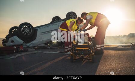 Sull'automobile Crash scena di incidente di traffico: Paramedics che salva la vita di una vittima femminile che sta mentendo su Stretchers. Ascoltano un Heartbeat, si applicano Foto Stock