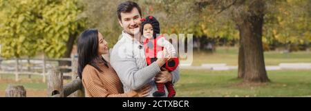 Felice sorridente madre cinese asiatica e padre caucasico papà con bambina in costume ladybug. Famiglia in autunno parco all'aperto. Halloween Foto Stock