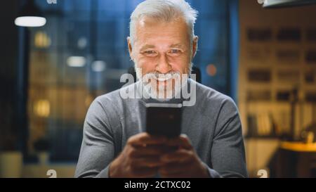 Ritratto del bell'uomo d'affari di media età Bearded usa lo smartphone mentre si siede alla sua scrivania, ride e sorride. Lavoro da Foto Stock