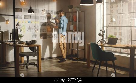 Young Creative Man si trova accanto al muro con la mood Board organizzata nel suo Cool Loft Office. Prende appunti su un pezzo di carta. Giornata di sole all'aperto Foto Stock