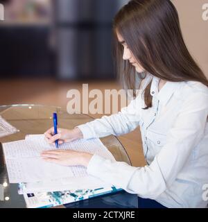 Ritratto quadrato della ragazza bella seria giovane con i capelli lunghi biondi con il suo lavoro su sfondo sfocato. Studentessa è seduta al tavolo Foto Stock