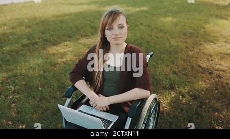 Espressione facciale di giovane disabile caucasica in sedia a rotelle con computer portatile nel parco. Foto di alta qualità Foto Stock