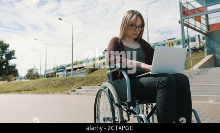 Giovane donna caucasica in sedia a rotelle utilizzando un computer portatile. Treno passando sullo sfondo. Foto di alta qualità Foto Stock