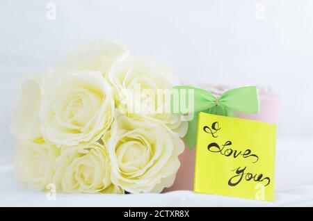 Regalo romantico. Bouquet di rose bianche, confezione regalo e nota "i Love You" posta su un letto. Primo piano. Foto Stock