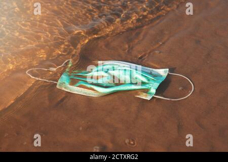 rifiuti infettivi - maschera medica viso in acqua sulla spiaggia di sabbia. mare e inquinamento oceanico dal covid-19 Foto Stock