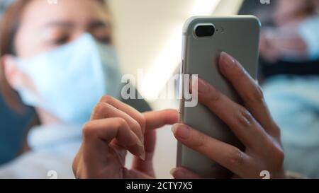 Donna asiatica che tiene lo smartphone e indossa una maschera contro le infezioni trasmissibili di 2019-nCoV durante il viaggio di volo. Viaggiatori che usano maschere per prote Foto Stock