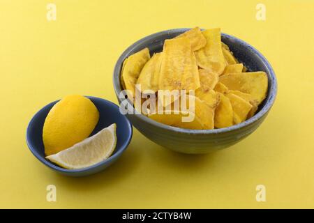 Patatine di plantain aromatizzate al limone in una ciotola per spuntini con spicchi di limone in ciotola blu su sfondo giallo Foto Stock