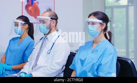 Stanco team di personale medico con maschera facciale e visiera contro l'epidemia di coronavirus nell'area di attesa dell'ospedale. Paziente che entra nella hall dell'ospedale. Stetoscopio medico. Foto Stock