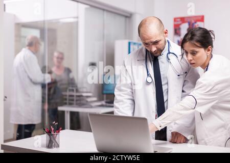 Team di giovani medici che lavorano insieme in ospedale per controllare l'evoluzione dei pazienti su un computer portatile. Donna anziana anziana e medica senior sul corridoio della clinica. Medico che indossa cappotto bianco e stetoscopio. Foto Stock