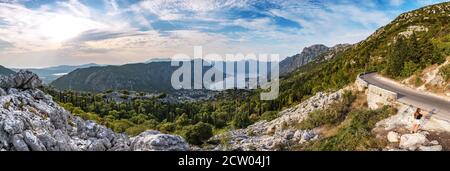 Incantevole panorama del Montenegro, città di Cattaro e baia dalla strada a serpentina di montagna. Belle montagne del Montenegro sopra il cielo con le nuvole al tramonto. Bella giovane donna che fa un selfie. Foto Stock