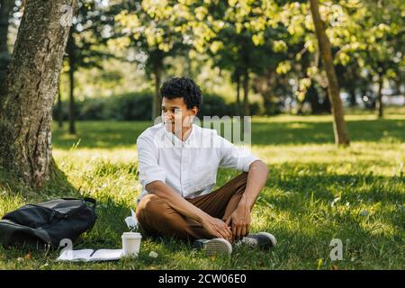 Giovane uomo sorridente seduto nel parco e guardando via Foto Stock