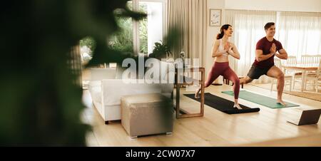 Uomo e donna che praticano lo yoga a casa con il computer portatile davanti. Coppia che guarda il video tutorial di fitness online sul computer portatile e facendo yoga. Foto Stock