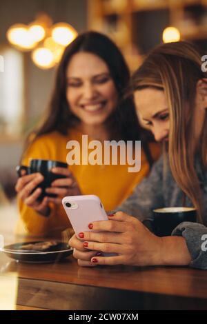 Due giovani donne che utilizzano uno smartphone presso la caffetteria. Due amiche sorridenti che usano un telefono cellulare in un bar Foto Stock