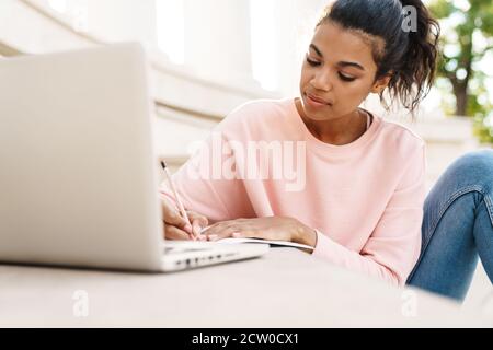 Immagine della ragazza studentesca afroamericana felice che fa il lavoro con computer portatile mentre si siede sulle scale all'aperto Foto Stock