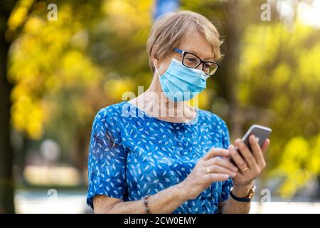 Donna anziana che usa lo smartphone mentre indossa una maschera protettiva per il viso Foto Stock