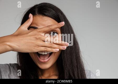 Eccitata giovane afroamericana indossando abiti casual in piedi isolati su sfondo grigio, copre faccia con le mani Foto Stock