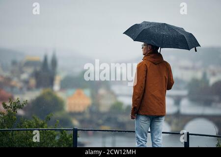 Giovane uomo con ombrello che guarda la città sotto la pioggia battente. Giornata cupa a Praga, Repubblica Ceca Foto Stock