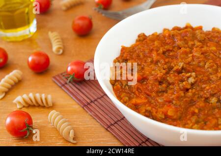 Ciotola piena di ragù classico italiano (salsa bolognese) sul tavolo con un po' di pasta, pomodori e un bicchiere di olio d'oliva Foto Stock