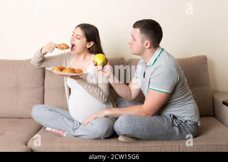 Marito cerca di nutrire la moglie incinta con una mela, ma vuole un croissant seduto sul divano di casa. Dieta non sana durante il concetto di gravidanza. Foto Stock