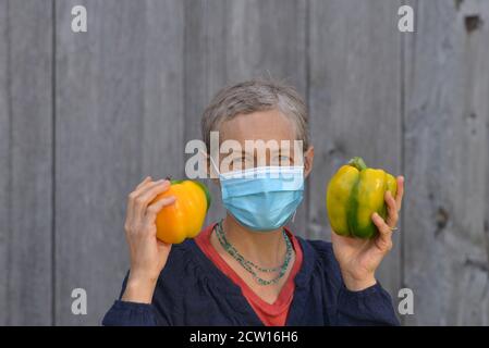 La donna caucasica canadese di medio-età con i capelli corti indossa una maschera chirurgica blu e tiene due peperoni organici grandi della campana nelle sue mani. Foto Stock