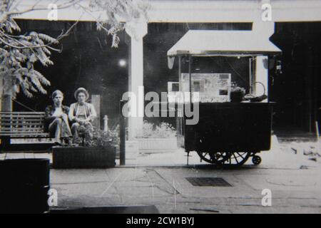 Bella fotografia in bianco e nero degli anni '70 di due ragazzi appesi su un marciapiede della città e seduti su una panchina del parco. Foto Stock